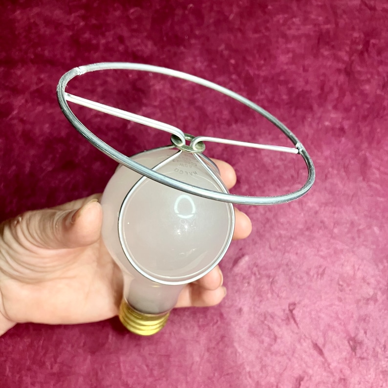 May include: A white light bulb with a silver metal clip top  lampshade ring around it. The ring is attached to the bulb with circular brass wires.