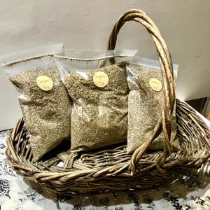 May include: A woven wicker basket holds three clear plastic bags filled with dried oregano. Each bag has a small, round label that reads "Oregano 7g". The basket has a curved handle and sits on a floral patterned surface.