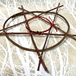 Könnte beinhalten: Ein handgefertigtes Pentagramm aus Zweigen und Ästen. In der Mitte befindet sich ein roter, gewebter Kreis mit roten Fadenakzenten. Das Design ist vor einem weißen und grauen Hintergrund angeordnet.