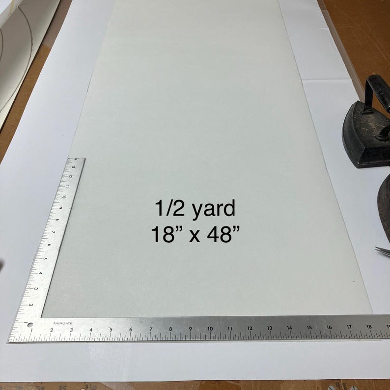 May include: A rectangular piece of solid white fabric positioned next to a metal L-shaped ruler. The fabric has the text 1/2 yard 18 inches by 48 inches printed in the center. A black iron is visible in the upper right corner of the frame.