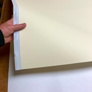 May include: A hand holding a large, off-white sheet of paper with a white adhesive strip along the edge. The paper is smooth and appears to be a print or art material.