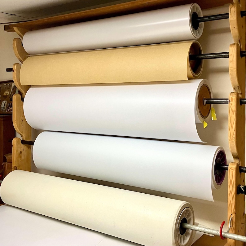 5 rolls of different kinds of lampshade styrene on a cutting rack. Rolls are 48” wide and hold various colors of adhesive and non adhesive styrene like white, cream and tan.