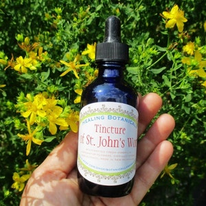 Könnte beinhalten: Eine blaue Glasflasche mit Tropfer und einem weißen Etikett mit der Aufschrift "Healing Botanicals Tincture of St. John's Wort". Die Flasche wird in einer Hand vor einem Hintergrund aus gelben Blumen gehalten.