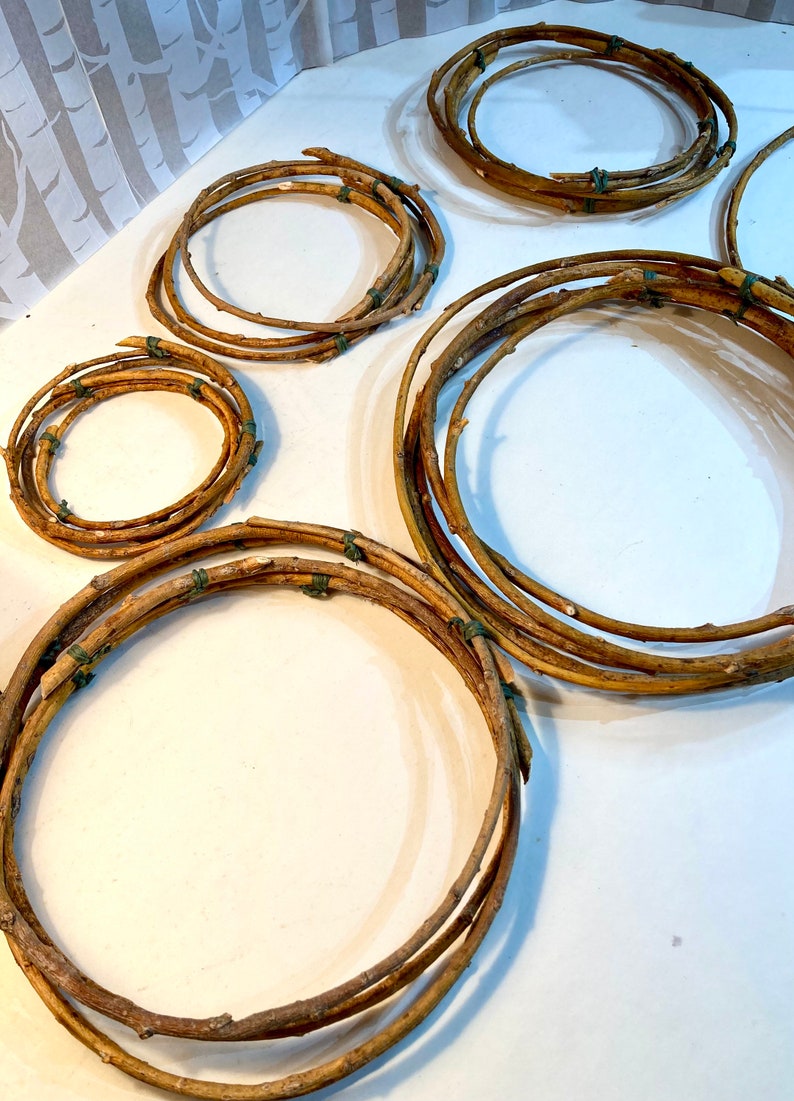 May include: Five different sized wreaths made of natural brown twigs tied together with green twine. The wreaths are arranged on a white surface.