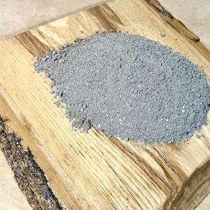 May include: A close-up of a pile of gray, fine-grained powder on a wooden surface.