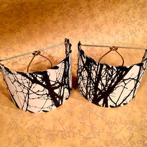 May include: Two white lampshades with a black tree branch pattern. The lampshades are attached to a gold metal frame.