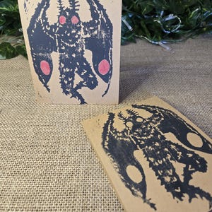 May include: Two brown cards with black ink illustrations of a creature with wings and red circles. The creature has a large head with a pointed beak and a long, thin body.