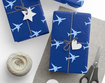 May include: Dark blue wrapping paper featuring a repeating pattern of light blue airplanes. The packages are tied with brown twine and decorated with white paper gift tags in the shapes of a star and a heart, accompanied by scissors and twine.