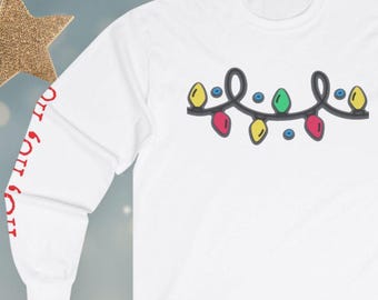May include: A white long-sleeve shirt featuring a graphic of a string of colorful Christmas lights in yellow, green, and red. The left sleeve displays the red text HO HO HO printed vertically along the arm, creating a festive holiday design.