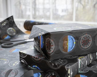 May include: Black wrapping paper featuring a repeating pattern of aviation instrument gauges. Two wrapped gifts are stacked, with a roll of paper and scissors visible. A gray ribbon is curled on the surface. The gauges have white, red, blue, and brown details.
