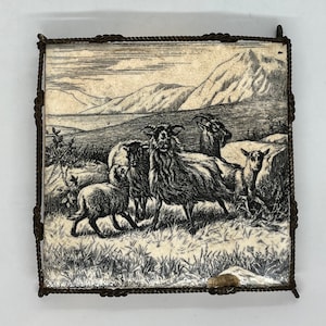 May include: A framed black and white etching of sheep and goats in a landscape with mountains and a body of water. The frame is a dark, twisted metal. The etching is on a cream-colored surface.