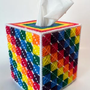 May include: A square tissue box cover crafted from plastic canvas with a vibrant rainbow-colored diagonal grid pattern. The edges are stitched with white yarn, and the top features a central opening with a rainbow border holding a white tissue.