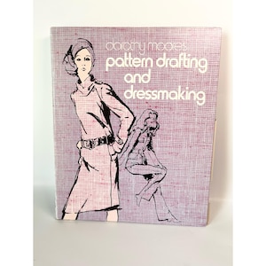 May include: A hardcover book titled Pattern Drafting and Dressmaking by Dorothy Moore. The cover features a light purple background with a dark purple grid pattern and black line illustrations of two figures wearing clothing designs.