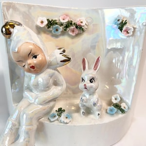 May include: A pearlescent white ceramic planter featuring a seated figure with a gold-accented hat and a small white rabbit. The planter is decorated with small floral accents in pink and blue.