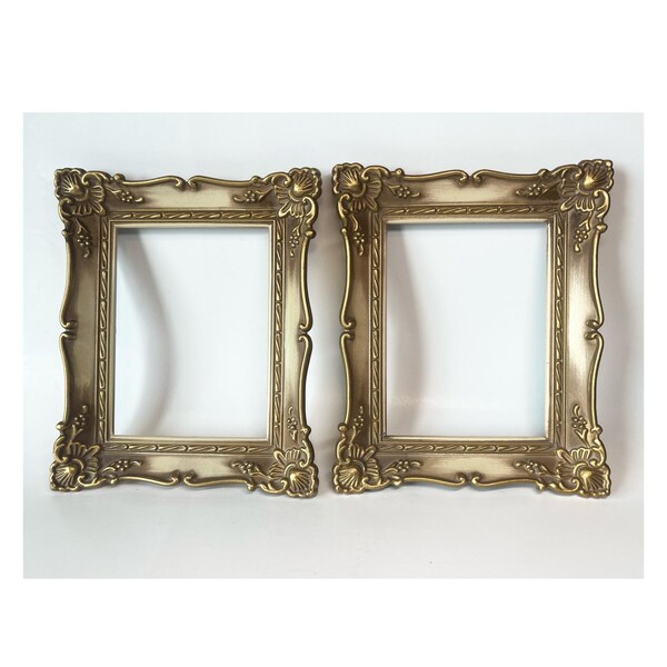 May include: Two ornate, rectangular picture frames with a gold finish and cream-colored accents. The frames feature detailed floral and scrollwork designs, perfect for displaying photos or artwork. The frames are empty.
