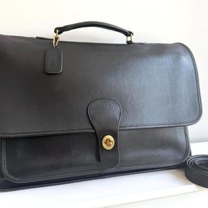 May include: A dark brown leather briefcase featuring a top handle, a fold-over flap with a brass turn-lock closure, and a matching leather luggage tag attached to the handle. A coiled, detachable leather shoulder strap lies to the right side.