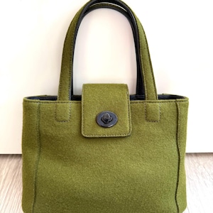 May include: An olive green handbag with black handles and trim. The bag has a rectangular shape with a flap closure and a circular clasp. The arched handles and the body of the bag appear to be made of a felt-like material.