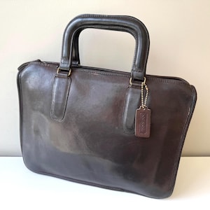 Può includere: Una borsa in pelle marrone scuro con manico superiore e forma rettangolare. La borsa ha una chiusura con cerniera e un piccolo tag in pelle con una catena attaccata. La borsa è su una superficie bianca.