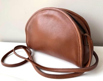 May include: A brown leather crossbody bag with a half-moon shape. The bag has a long, thin strap and a black zipper. The leather appears to be smooth and has a slight sheen. The bag is a classic style.
