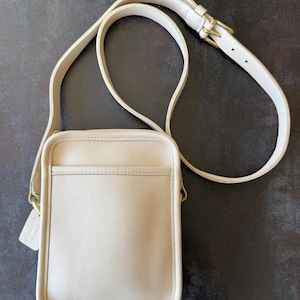 Könnte beinhalten: Eine cremefarbene Umhängetasche aus Leder mit rechteckiger Silhouette, einer vorderen Einstecktasche und einem verstellbaren Schulterriemen mit goldfarbenen Metallbeschlägen. Ein passender Lederanhänger ist an der Seite befestigt.
