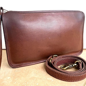 Könnte beinhalten: Eine braune Leder-Clutch mit Handschlaufe und abnehmbarem Schulterriemen. Die rechteckige Tasche hat einen Reißverschluss und messingfarbene Beschläge. Das Leder scheint abgenutzt zu sein und hat eine natürliche Maserung.