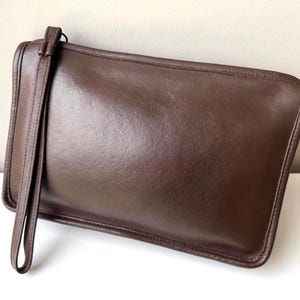 Op de afbeelding: Een rechthoekige, bruine leren clutch met een polsbandje. De tas heeft een gladde, glanzende afwerking en een gestikte rand. Het polsbandje is aan de bovenkant van de tas bevestigd. De clutch ligt op een wit oppervlak.