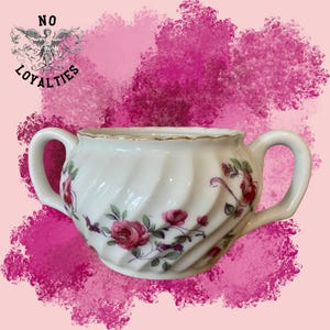 Puede incluir: Azucarero de porcelana blanca con un diseño de remolino y un patrón floral de rosas rosas. El tazón tiene dos asas y un borde dorado.