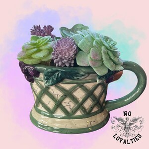 Könnte beinhalten: Ein grüner Keramik-Pflanztopf in Form einer Teetasse mit Henkel, gefüllt mit künstlichen Sukkulenten in Grün- und Lilatönen.