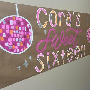 May include: A brown paper banner with the text "Cora's Sweet Sixteen" in pink, orange, and white lettering. The banner is decorated with two silver glitter disco balls.