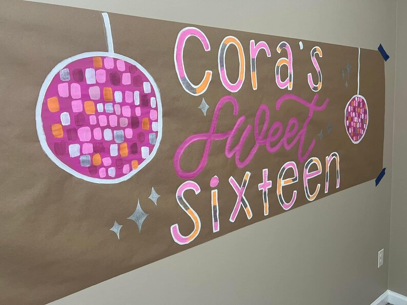 May include: A brown paper banner with the text "Cora's Sweet Sixteen" in pink, orange, and white lettering. The banner is decorated with two silver glitter disco balls.