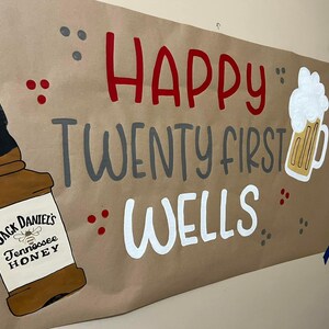 May include: A brown paper banner with the text "Happy Twenty First Wells" in red, gray, and white paint. A bottle of Jack Daniel's Tennessee Honey whiskey and a glass of beer are painted on the banner.