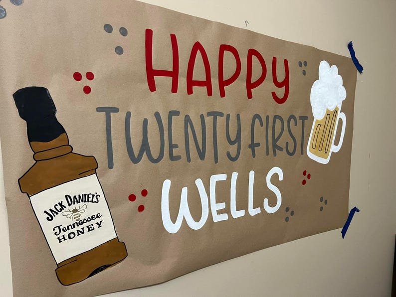May include: A brown paper banner with the text "Happy Twenty First Wells" in red, gray, and white paint. A bottle of Jack Daniel's Tennessee Honey whiskey and a glass of beer are painted on the banner.