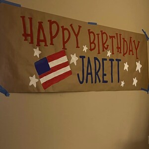 May include: A brown paper banner with the words "Happy Birthday Jarett" in red and blue paint. The banner also features a white star and a red, white, and blue American flag.