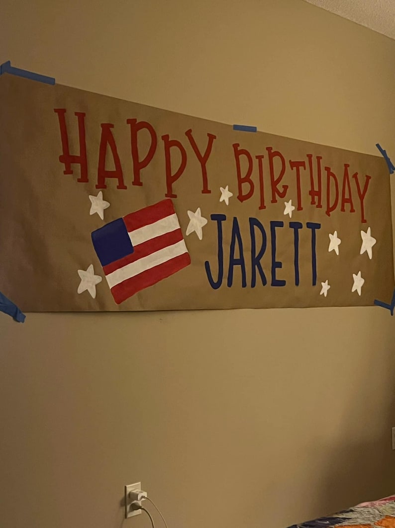 May include: A brown paper banner with the words "Happy Birthday Jarett" in red and blue paint. The banner also features a white star and a red, white, and blue American flag.
