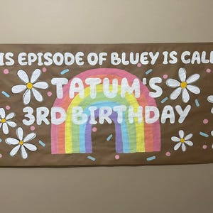 May include: A brown paper banner with the text "This episode of Bluey is called Tatum's 3rd Birthday" in white paint. The banner is decorated with a rainbow, white daisies, and pink and blue confetti.