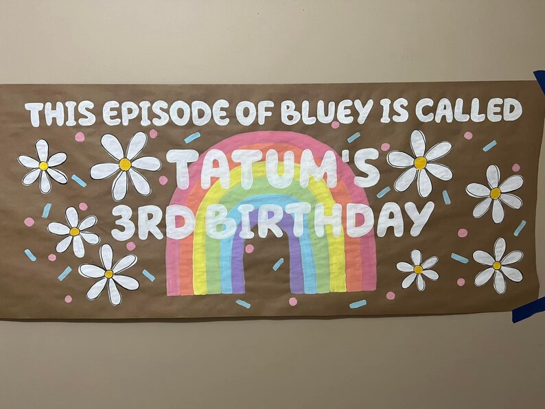 May include: A brown paper banner with the text "This episode of Bluey is called Tatum's 3rd Birthday" in white paint. The banner is decorated with a rainbow, white daisies, and pink and blue confetti.