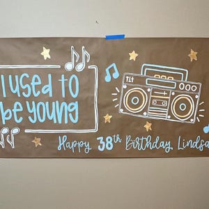 May include: A brown paper banner with the text "I used to be young" in white and blue lettering. A gold star and music notes are drawn on the banner. A cartoon boombox is drawn in the center of the banner with the text "Happy 38th Birthday Lindsay" in white and blue lettering.