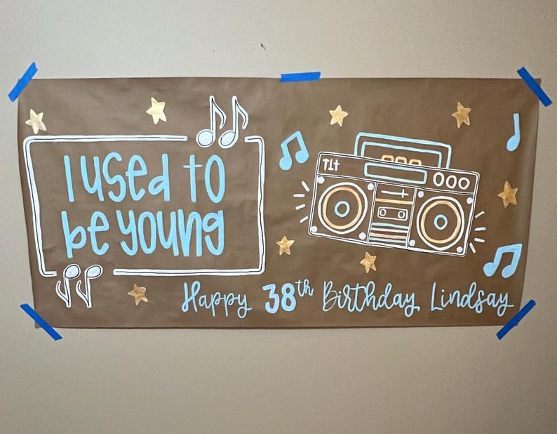 May include: A brown paper banner with the text "I used to be young" in white and blue lettering. A gold star and music notes are drawn on the banner. A cartoon boombox is drawn in the center of the banner with the text "Happy 38th Birthday Lindsay" in white and blue lettering.
