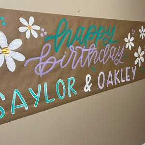 May include: A brown paper banner with the words "Happy Birthday Saylor & Oakley" written in purple and teal paint. The banner is decorated with white daisies.