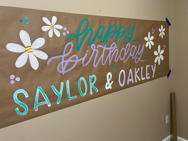 May include: A brown paper banner with the words "Happy Birthday Saylor & Oakley" written in purple and teal paint. The banner is decorated with white daisies.