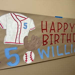 May include: A brown paper banner with the words "Happy 5th Birthday William" in red and blue paint. The banner also features a white baseball jersey with red trim and the number 5, a brown baseball glove, a brown baseball bat, and a white baseball with red stitching.