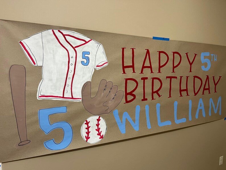 May include: A brown paper banner with the words "Happy 5th Birthday William" in red and blue paint. The banner also features a white baseball jersey with red trim and the number 5, a brown baseball glove, a brown baseball bat, and a white baseball with red stitching.