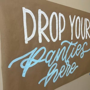 May include: A brown paper sign with the text "DROP YOUR PANTIES HERE" written in white and light blue paint.