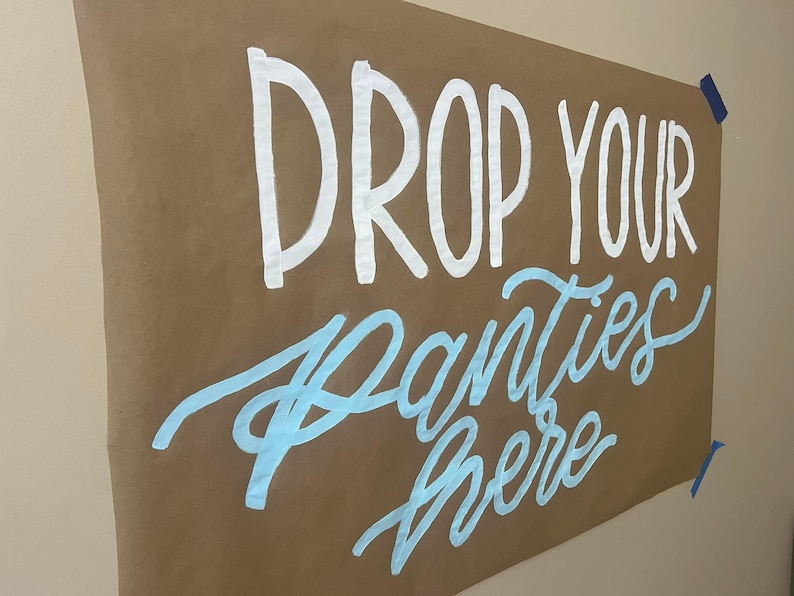 May include: A brown paper sign with the text "DROP YOUR PANTIES HERE" written in white and light blue paint.