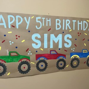 May include: A brown paper banner with the text "Happy 5th Birthday Sims" in blue paint. The banner is decorated with red, yellow, and blue confetti. Three monster trucks are painted on the banner in green, red, and blue.