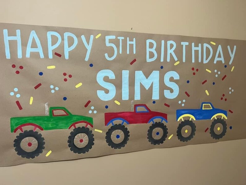 May include: A brown paper banner with the text "Happy 5th Birthday Sims" in blue paint. The banner is decorated with red, yellow, and blue confetti. Three monster trucks are painted on the banner in green, red, and blue.