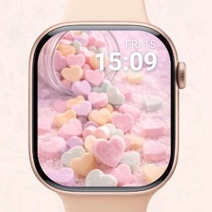 Op de afbeelding: Een gouden Apple Watch toont een scherm met een digitale klok die 15:09 uur en de datum weergeeft. De wijzerplaat toont een omgevallen pot met pastelkleurige hartvormige snoepjes, waarvan er één "LOVE YOU" leest. De horlogeband is een bijpassend lichtroze.