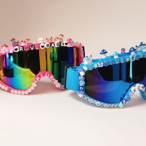 Könnte beinhalten: Zwei Paar Skibrillen, die mit bunten Perlen und Charms verziert sind. Die Brille links ist pink mit Regenbogenlinsen und dem Text "RAVE BABE PLUR" auf dem Rahmen. Die Brille rechts ist blau mit blauen Linsen und dem Text "RAVE PLUR EDM RAILRIDER" auf dem Rahmen.