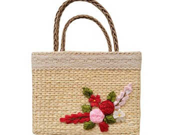 May include: A rectangular straw tote bag with braided handles and a cream-colored lace trim. The bag is adorned with a floral arrangement of red, pink, and white fabric flowers and green leaves, adding a decorative touch to the natural-toned bag.