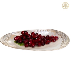 May include: An oval-shaped mother-of-pearl dish featuring ornate silver-toned metal accents on both ends, filled with a cluster of deep red, glossy artificial grapes with a small green leaf tucked underneath the fruit arrangement.